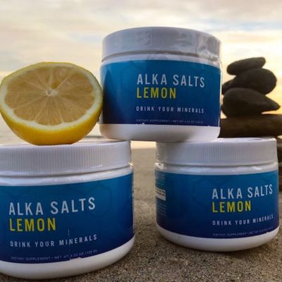 Three jars of Alka Salts Lemon stacked on a beach at dusk with a lemon half